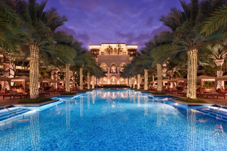 Piscina exterior iluminada del Palace Downtown con palmeras decoradas y arquitectura árabe al atardecer