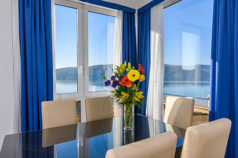 Comedor con mesa de cristal y vistas panorámicas al mar en Villa Mediterana con cortinas azules