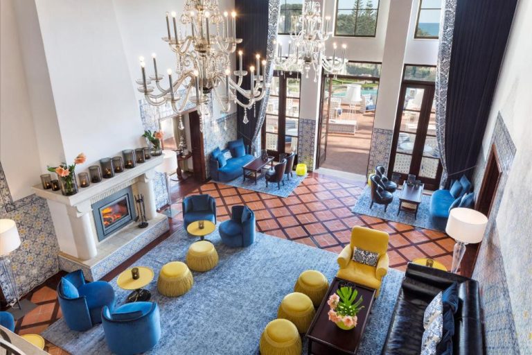 Elegante salón del Vila Vita Parc con chimenea, lámparas de cristal y sofás azules y amarillos