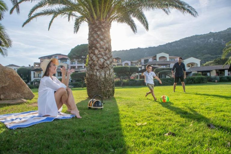 Familia jugando al fútbol en los jardines con palmeras del Hotel Abi d'Oru en Cerdeña