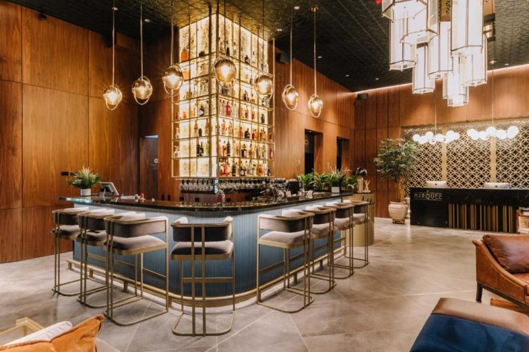 Bar lounge elegante con estantería de licores iluminada y taburetes dorados en Hotel Marquee Medellín