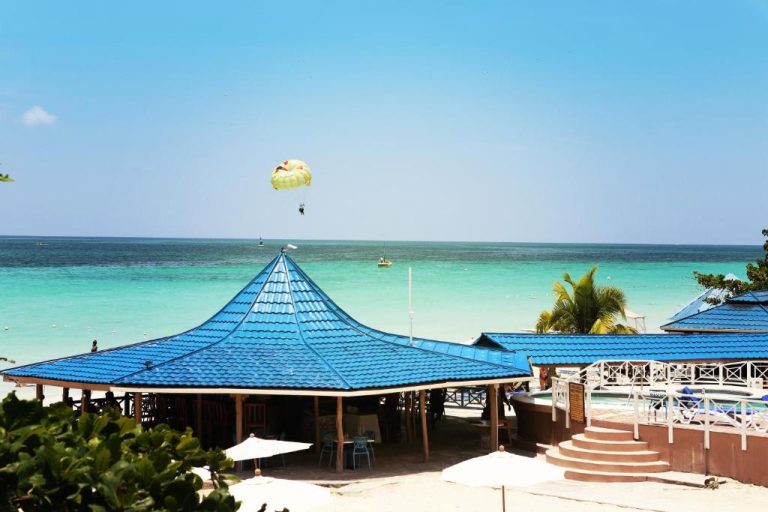 Vista de la playa turquesa con parasailing y pabellón azul del Negril Treehouse Resort en Jamaica