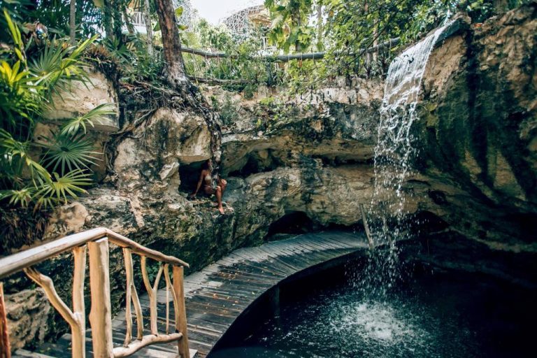 Cenote natural con cascada y pasarela de madera rodeado de selva tropical en Kan Tulum Hotel
