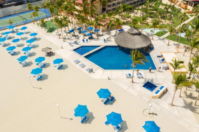 Vista aérea de la piscina con palapa y playa del hotel Posada Real Los Cabos con tumbonas azules