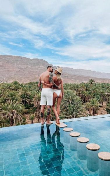Pareja romántica en la piscina infinita del Kasbah Hnini con vistas al oasis de palmeras y montañas