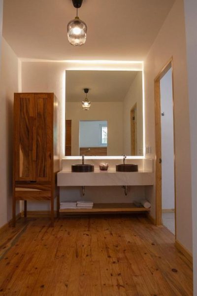 Baño doble con lavabos de piedra, espejo retroiluminado y suelo de madera en NaNa Vida CDMX