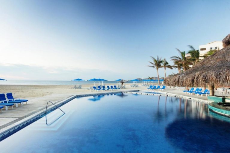 Piscina infinita frente a la playa del hotel Posada Real Los Cabos con tumbonas azules y palmeras