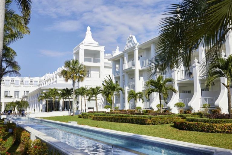 Fachada exterior blanca del Riu Palace Riviera Maya con piscina lineal y jardines tropicales