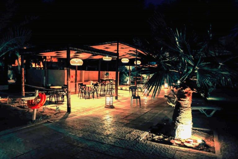 Terraza exterior iluminada del Hotel Continental con mesas, sillas y palmeras decorativas por la noche