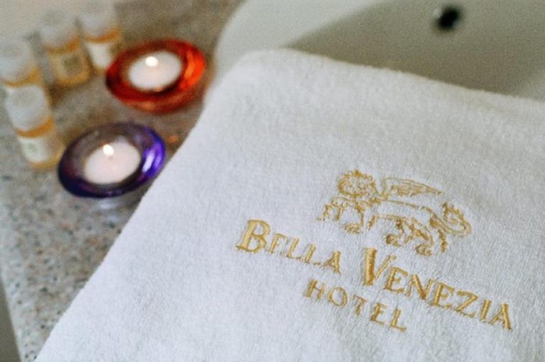 Toalla bordada con logo del Hotel Bella Venezia junto a velas y amenities de baño