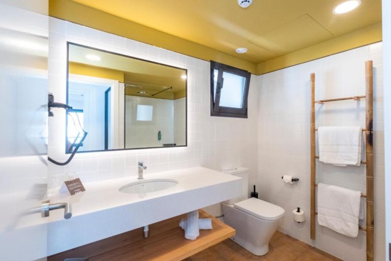 Baño moderno con lavabo doble, espejo iluminado y toallero de bambú en Calallonga Hotel Menorca