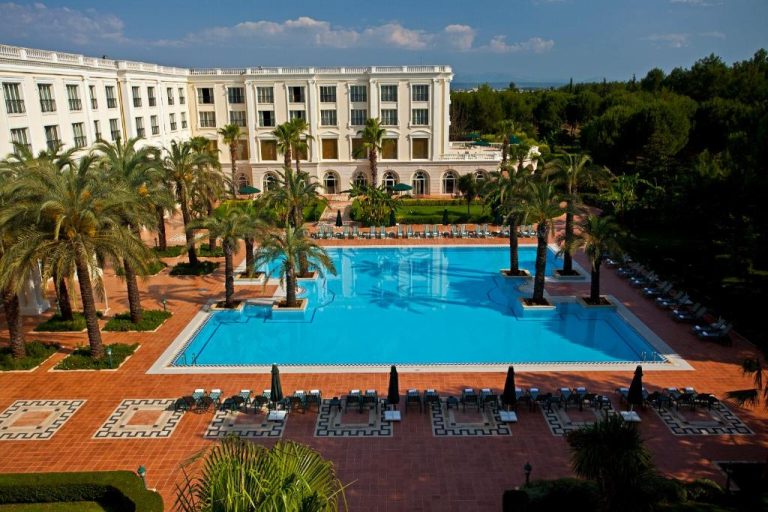 Piscina exterior rodeada de palmeras y tumbonas del IC Hotels Airport con fachada blanca al fondo