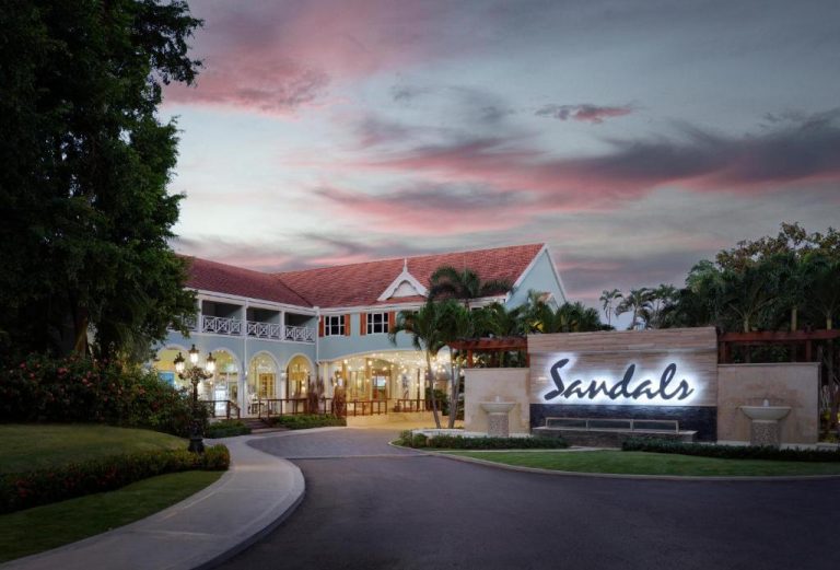 Entrada principal iluminada del Sandals Ochi Beach Resort al atardecer, rodeada de palmeras y jardines tropicales