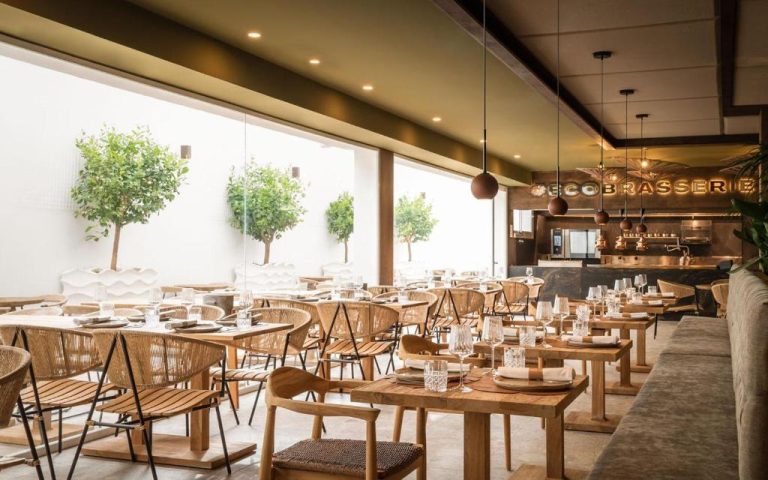 Restaurante EcoBrasserie del Lago Resort Menorca con mesas de madera, sillas rattan y árboles naturales interiores