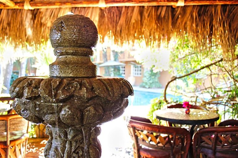 Fuente de piedra decorativa bajo palapa tropical con piscina al fondo en The Bungalows Hotel