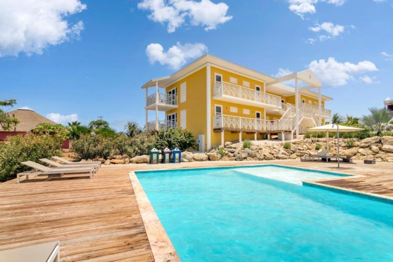 Piscina exterior con terraza de madera y villa amarilla en Chogogo Dive & Beach Resort Curaçao