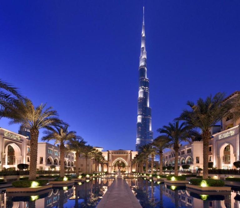 Vista nocturna del Palace Downtown Dubai con el Burj Khalifa iluminado y jardines con palmeras