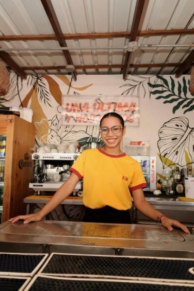 Camarera sonriente detrás de la barra del Bar Kim con decoración tropical y máquina de café