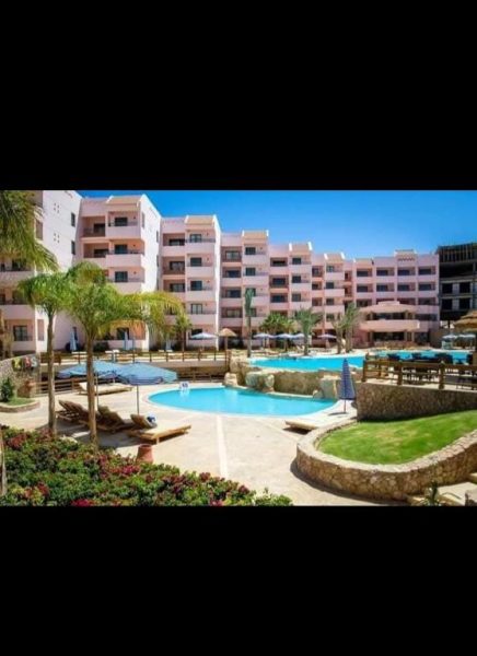 Piscina exterior con palmeras y tumbonas de la قرية الدهبية con edificios rosados al fondo