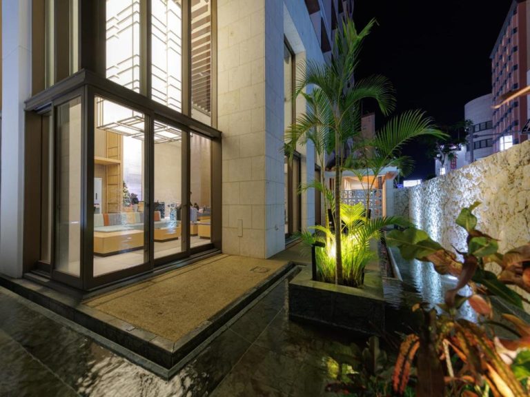 Entrada exterior iluminada del JR Kyushu Hotel Blossom Naha con jardín tropical y palmeras por la noche