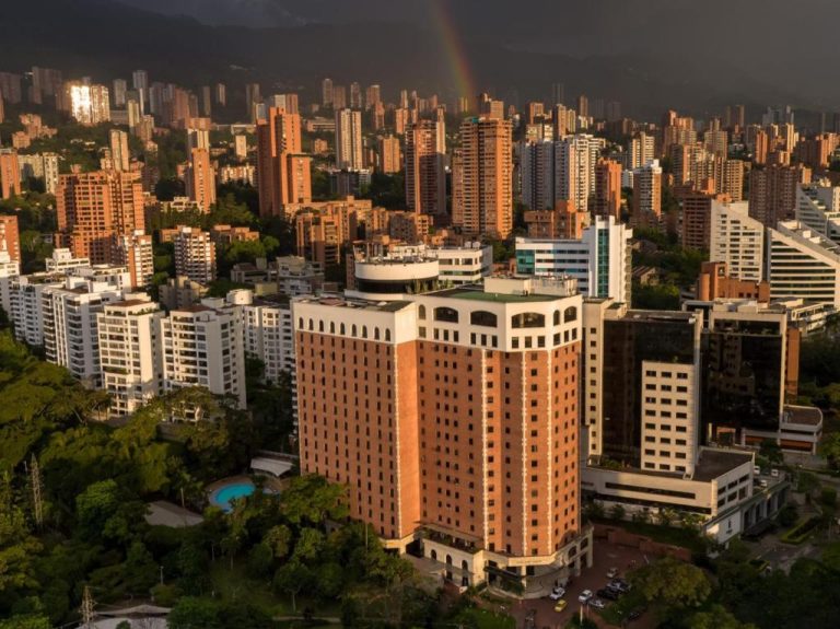 Vista aérea del Hotel Dann Carlton Medellín rodeado de edificios y vegetación con arcoíris al fondo