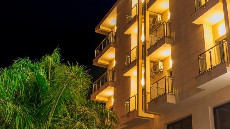 Fachada exterior iluminada del Hotel Rherssllah con balcones y palmeras durante la noche