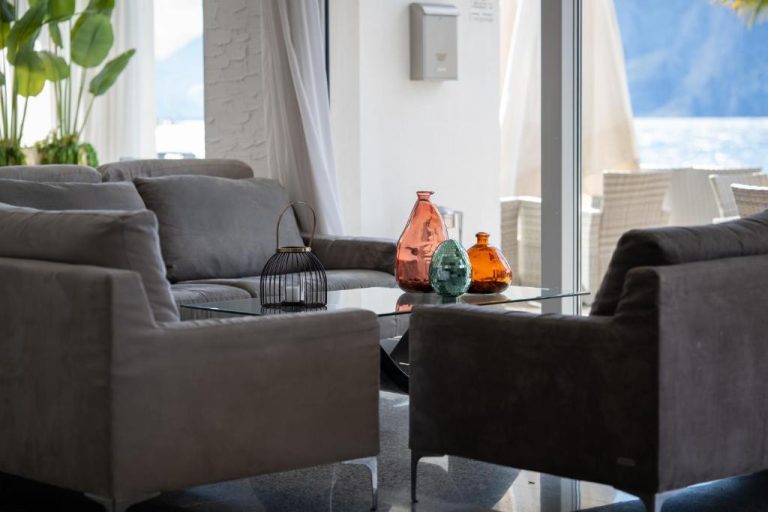 Elegante salón con sofás grises y mesa de cristal con decoración en Hotel Excelsior Bay con vistas al lago