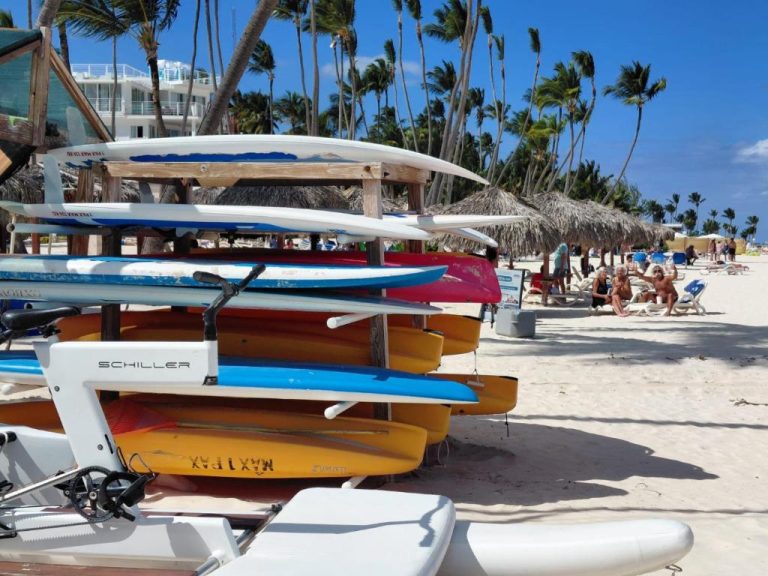Tablas de surf y kayaks en la playa del Hotel Punta Cana Coral Beach con palmeras y tumbonas al fondo