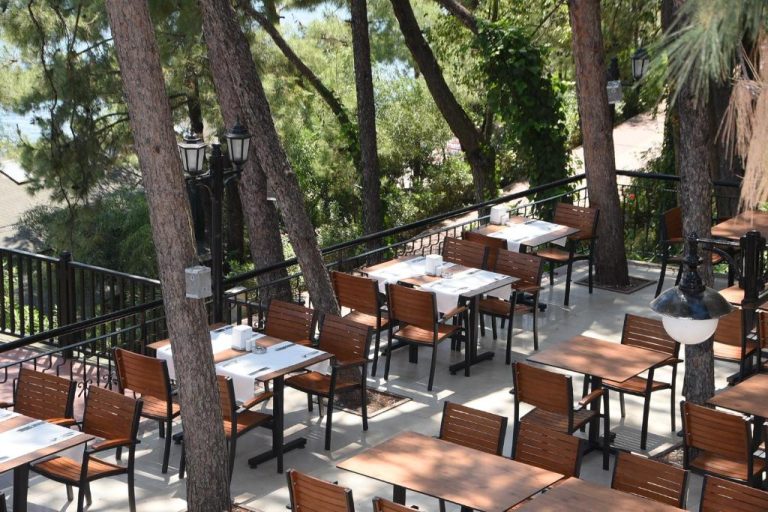 Terraza restaurante exterior con mesas y sillas de madera rodeada de pinos en Marmaris Park Hotel