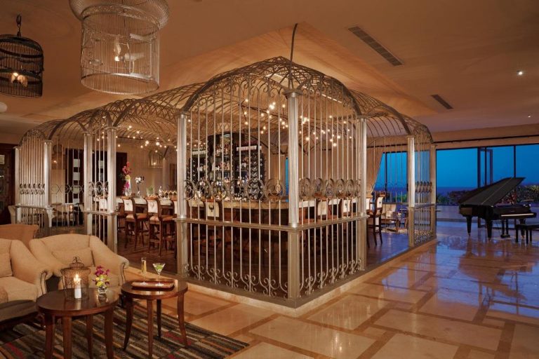 Bar interior con diseño de jaula metálica ornamental y piano de cola en Secrets Puerto Los Cabos Golf & Spa