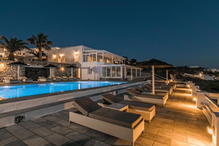Piscina exterior iluminada de noche con tumbonas y villa blanca del Hotel Alkyon en Mykonos