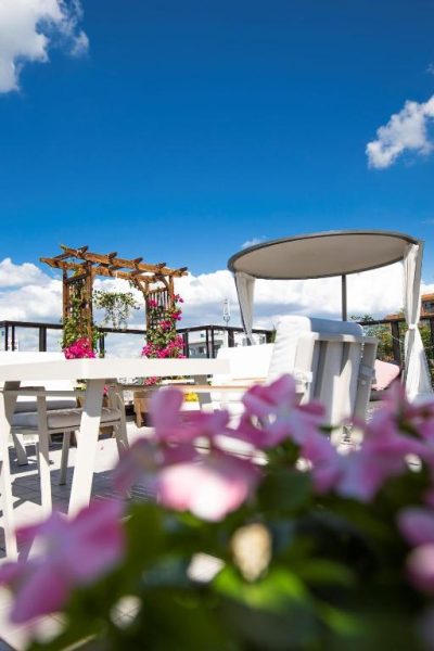 Terraza exterior con pérgola de madera, flores rosas y zona lounge del Sola Hotel bajo cielo azul