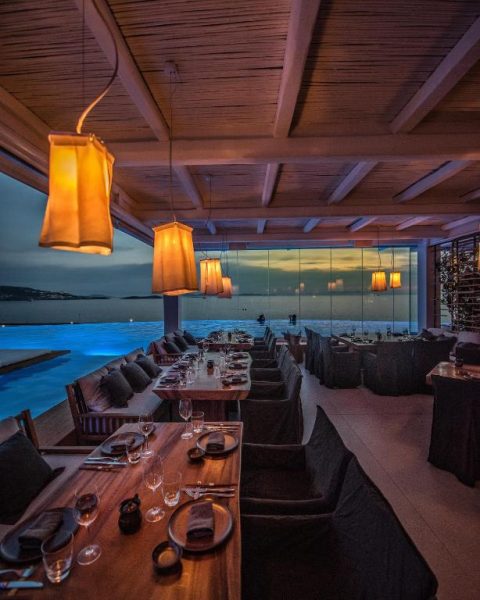 Restaurante con vista al mar al atardecer en Cavo Tagoo Mykonos con mesas de madera y lámparas cálidas