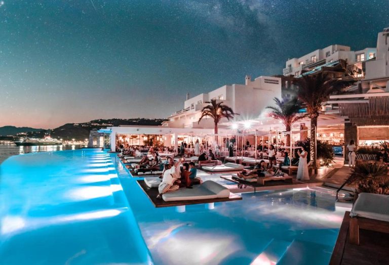 Piscina infinita iluminada de noche con tumbonas flotantes y vistas al mar en Cavo Tagoo Mykonos