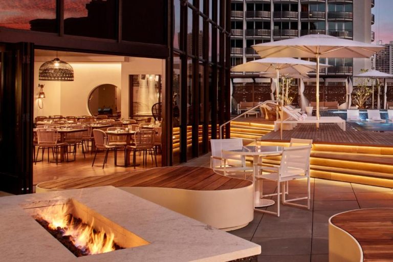 Terraza exterior con fogón, mesas y piscina iluminada al atardecer en Hyatt Centric Waikiki Beach