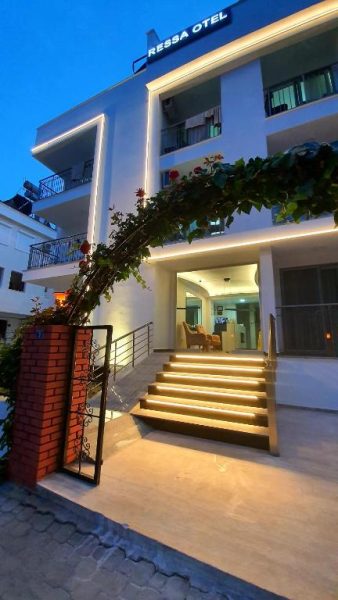 Entrada principal iluminada del Ressa Apart con escaleras y jardín en fachada blanca de noche