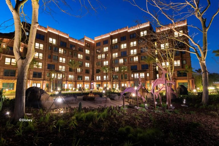 Fachada exterior iluminada del Disney Explorers Lodge con jardines y esculturas decorativas durante la noche