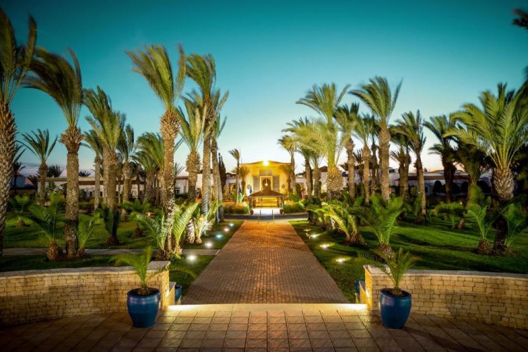 Jardines iluminados con palmeras y paseo central del hotel Robinson Agadir All Inclusive al atardecer