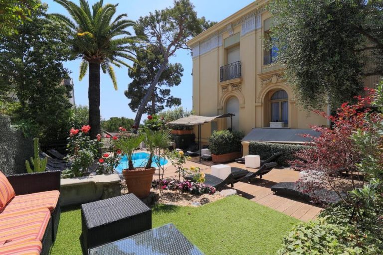 Jardín con piscina exterior y terraza del Hôtel Petit Palais rodeado de palmeras y vegetación mediterránea