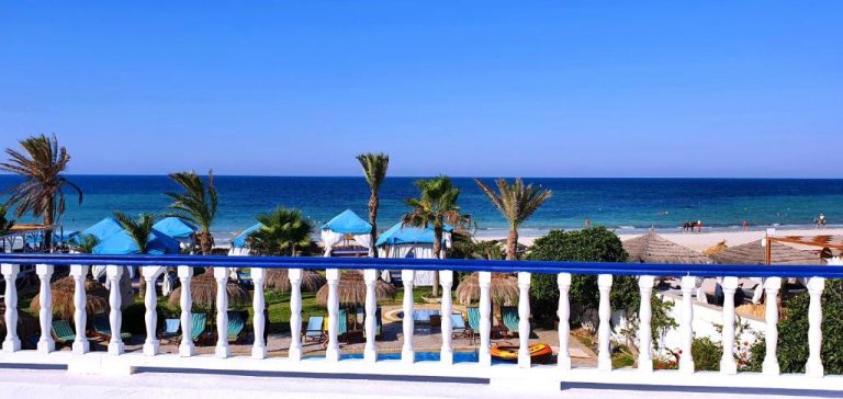 Terraza con balaustrada blanca y azul con vistas a la playa y palmeras del Hotel Dar El Bhar