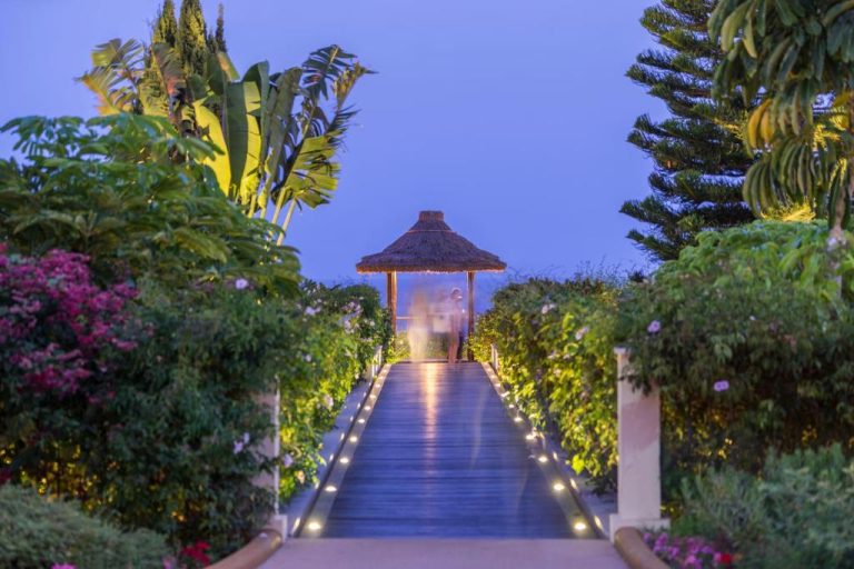 Pasarela iluminada con gazebo de paja rodeada de jardines tropicales al atardecer en Hotel Porto Mare PortoBay