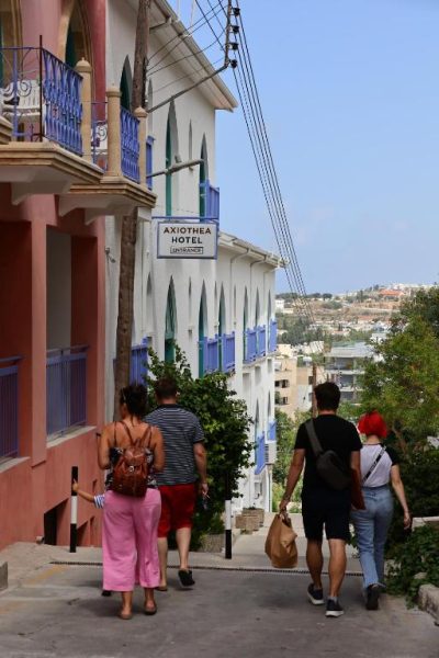 Turistas caminando por la calle de acceso a la entrada del Axiothea Hotel con fachada blanca y azul