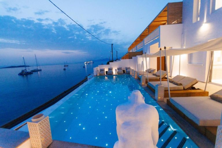 Piscina infinita iluminada con vistas al mar y veleros al atardecer en Icon Mykonos