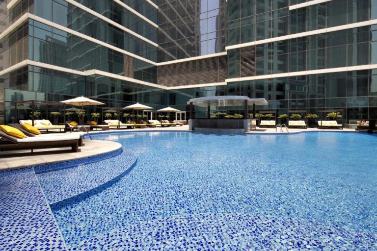 Piscina exterior con tumbonas y sombrillas del Taj Dubai rodeada de torres de cristal