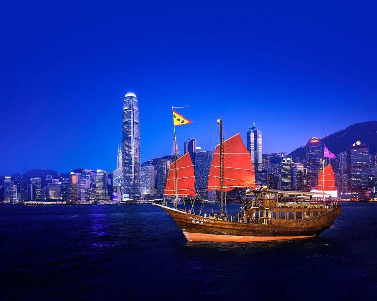 Barco junk tradicional con velas rojas navegando en Victoria Harbour frente al skyline del Four Seasons Hong Kong