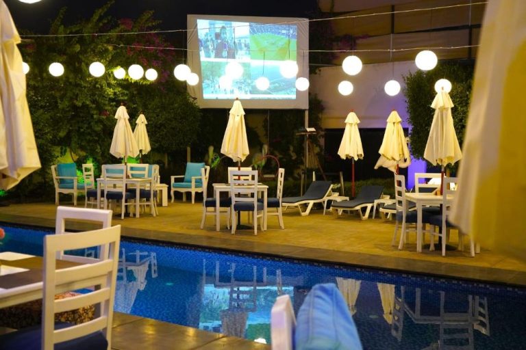 Piscina exterior iluminada de noche con tumbonas, sombrillas y pantalla de cine al aire libre en Aitch Boutique Hotel