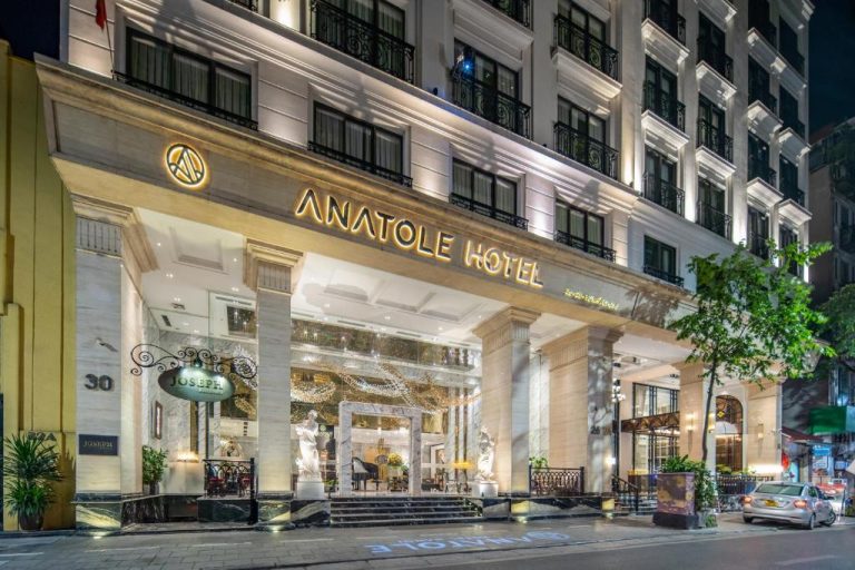 Fachada exterior iluminada del Anatole Hotel Hanoi con entrada principal y columnas de mármol de noche