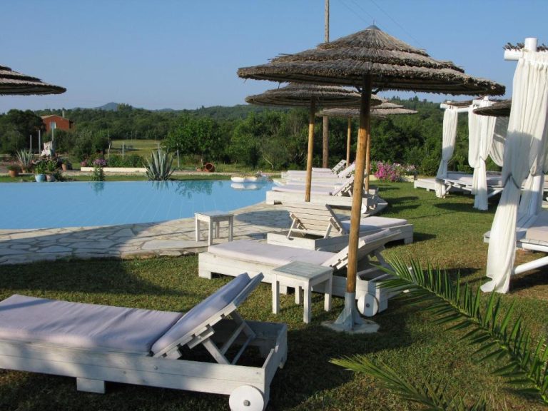 Piscina exterior con tumbonas blancas y sombrillas de paja en Pulades Resort rodeada de jardín
