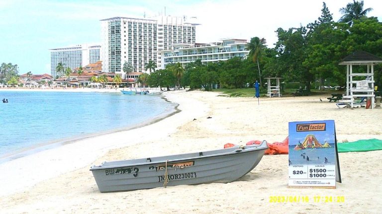 Playa tranquila con bote en arena y hoteles al fondo en Ocho Rios Vacation Resort, Jamaica