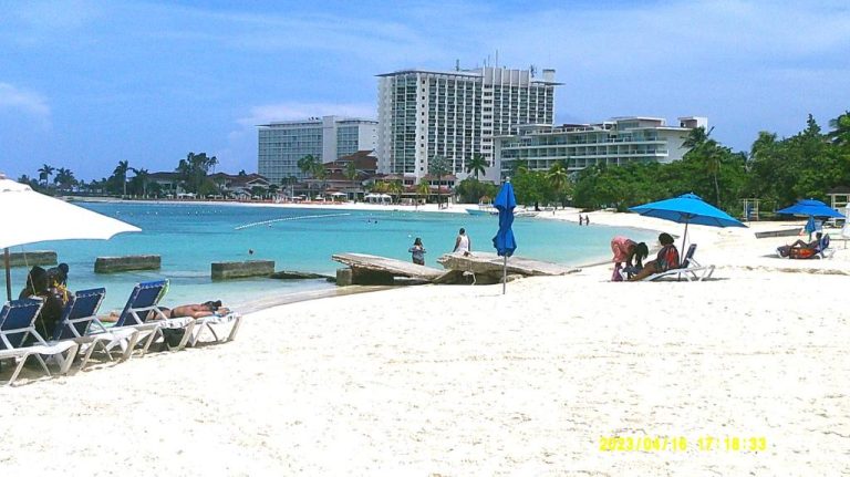 Playa de arena blanca con tumbonas y sombrillas azules frente al Ocho Rios Vacation Resort en Jamaica
